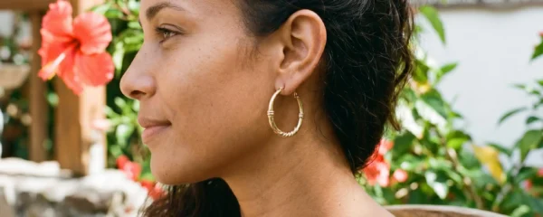 Gros plan sur une oreille portant des créoles dorées traditionnelles antillaises, avec des pétales d'hibiscus rouge en arrière-plan flou