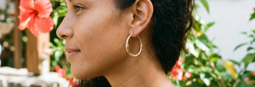 Gros plan sur une oreille portant des créoles dorées traditionnelles antillaises, avec des pétales d'hibiscus rouge en arrière-plan flou
