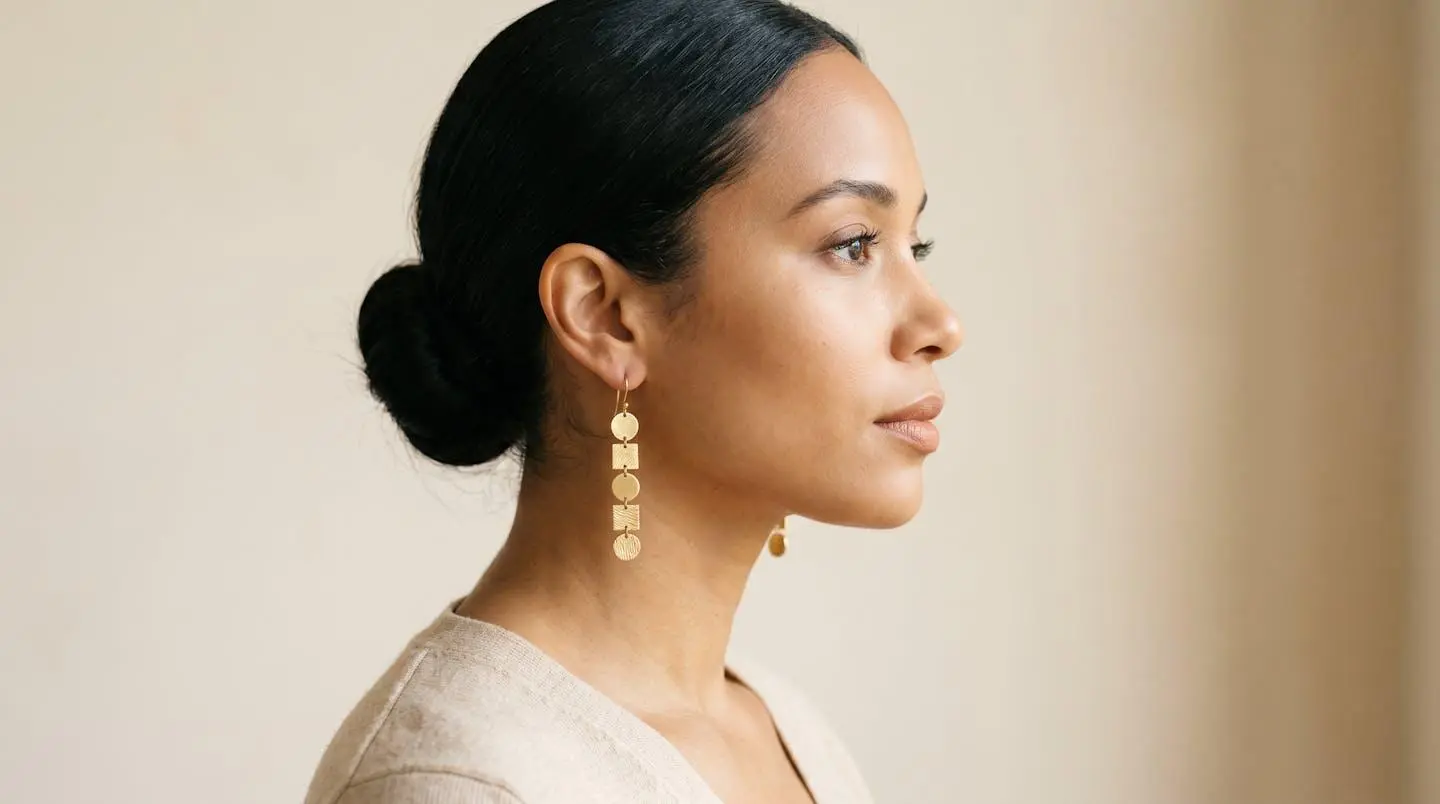 Portrait de profil d'une femme aux cheveux tirés en chignon portant des boucles d'oreilles pendantes dorées, sur fond beige épuré